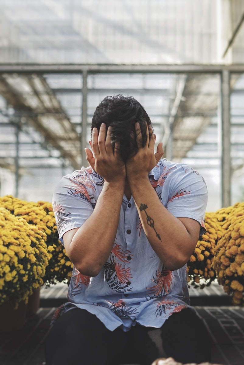 Man In Floral Shirt Covering His Face With His Hands Sitting Between Potted Yellow Flowers
