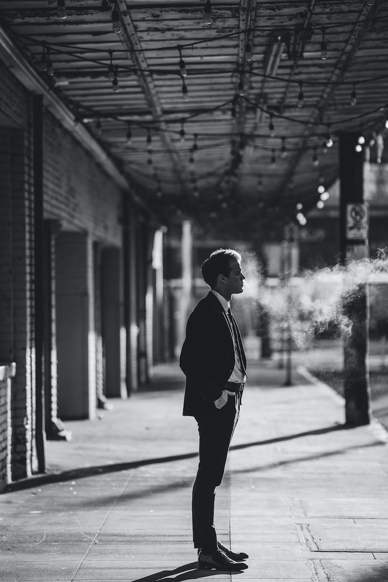 Black and white side view of male entrepreneur in formal suit with hand in pocket looking forward between haze and building on pathway