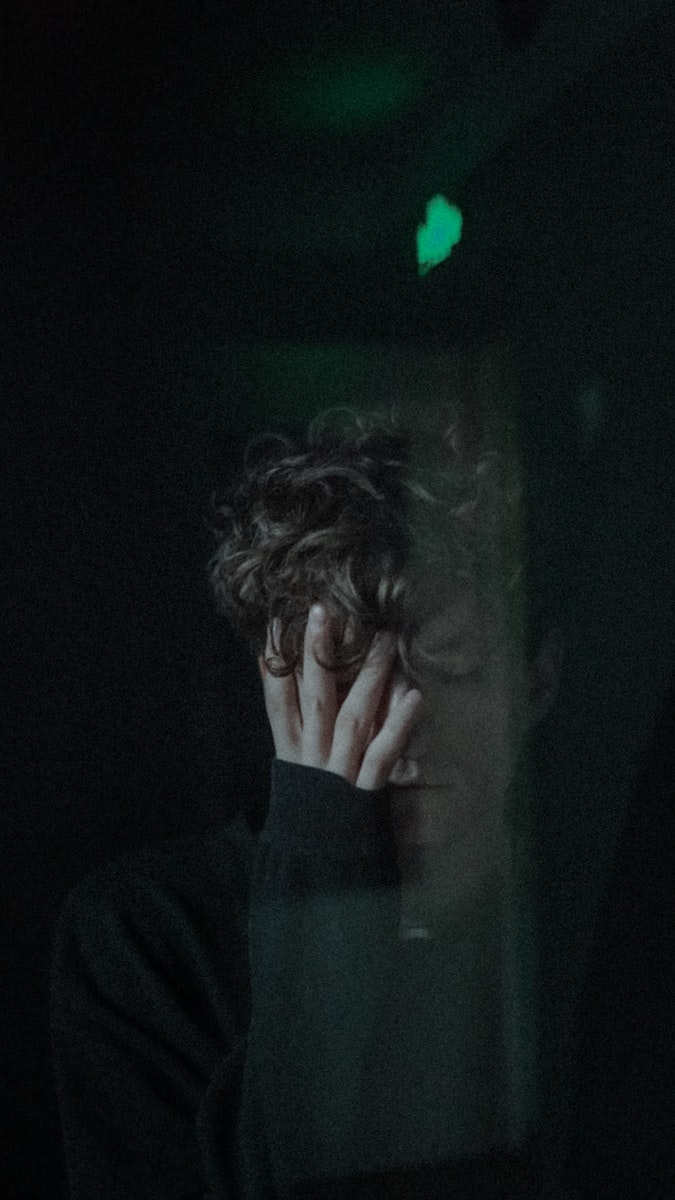 Through window of depressed unhappy male with closed eyes covering face with hand while standing in dark room near glass