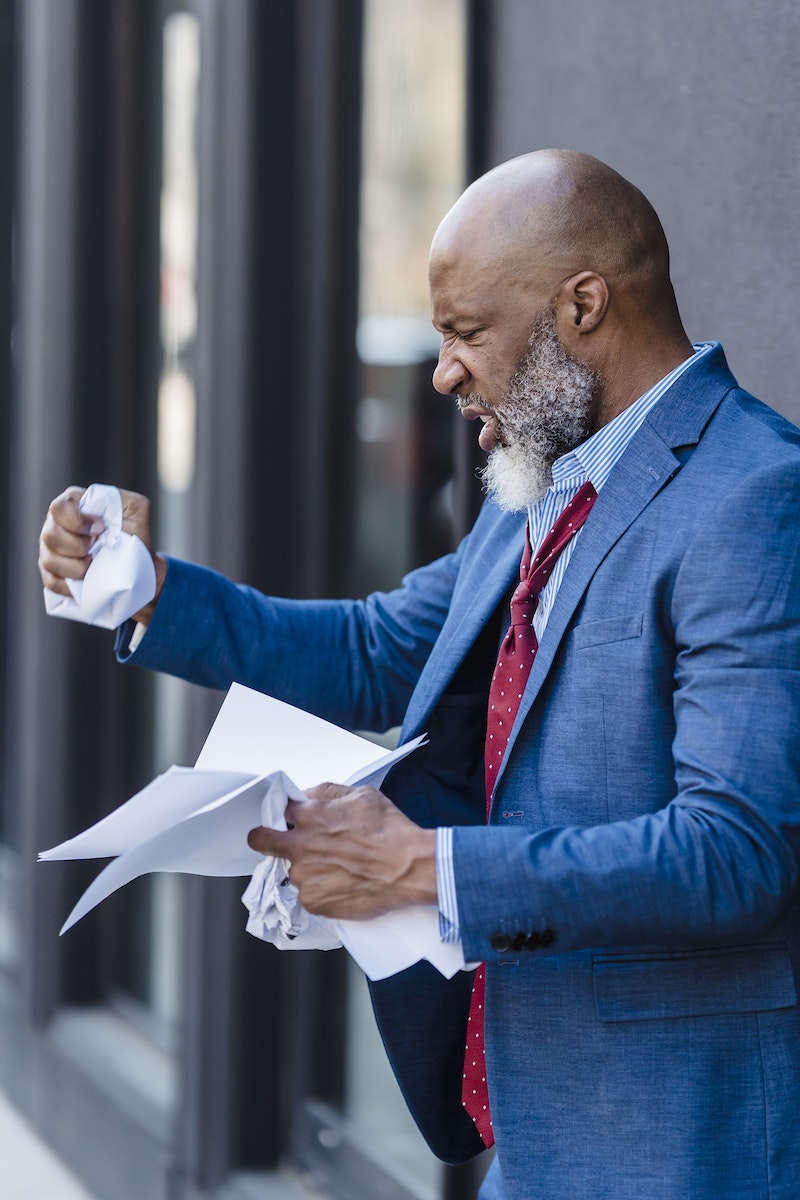 Side view of furious African American male entrepreneur in formal wear with documents in hands standing on street near building