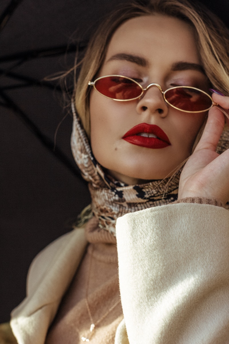 Low angle of gorgeous young lady with red lips in elegant outfit adjusting trendy sunglasses while standing on street with umbrella