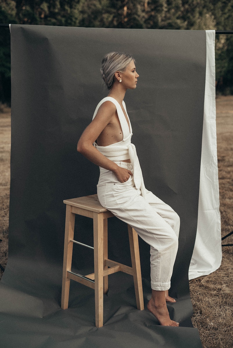 Side view of barefoot slim lady sitting on chair near gray background with hands in pockets in nature during photo shoot
