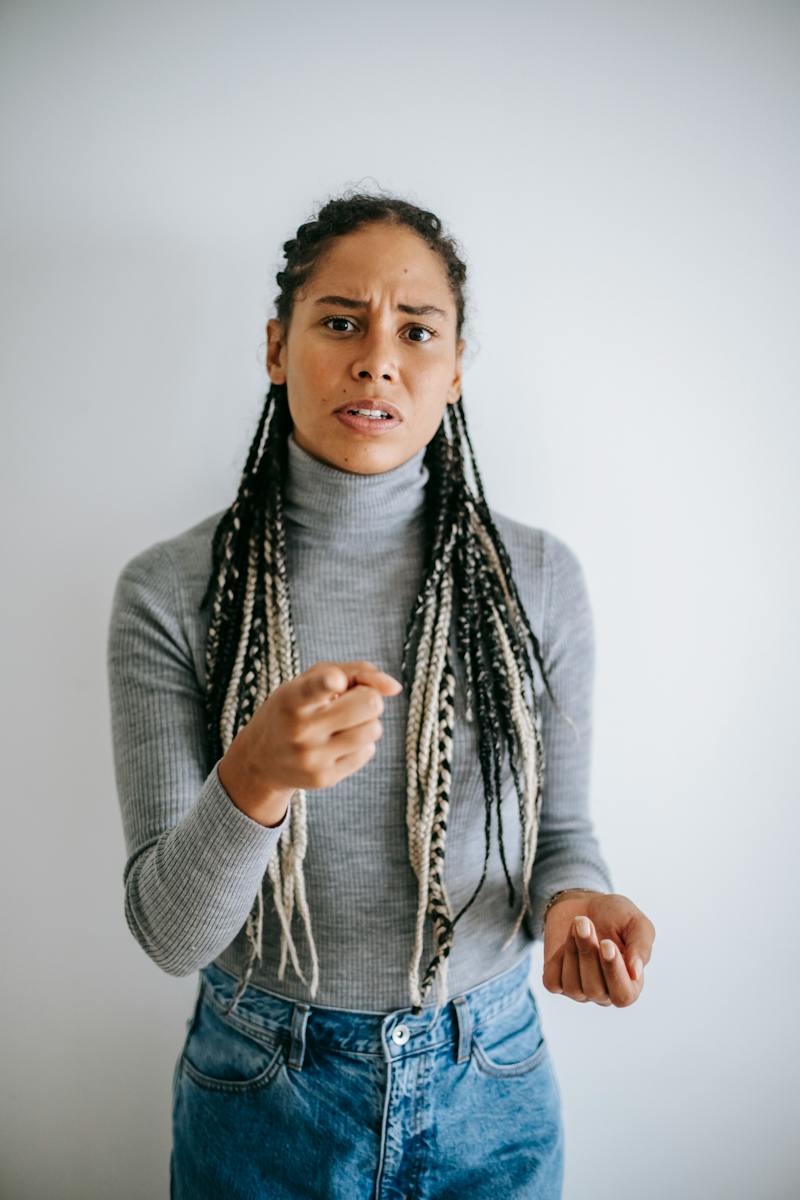 Ethnic female with long braids blaming pointing with forefinger at camera standing against gray background