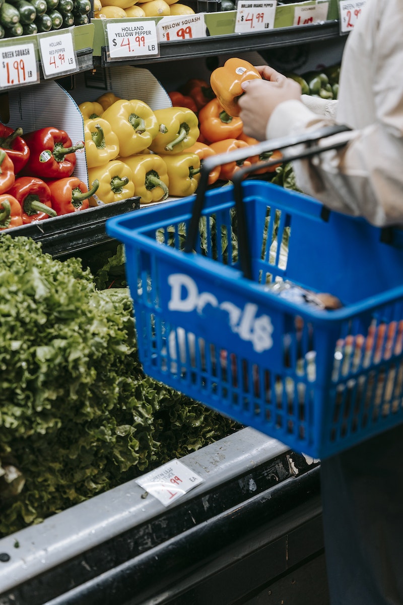 Side view of crop faceless buyer in casual clothes carrying shopping basket while choosing assorted colorful bell peppers in supermarket