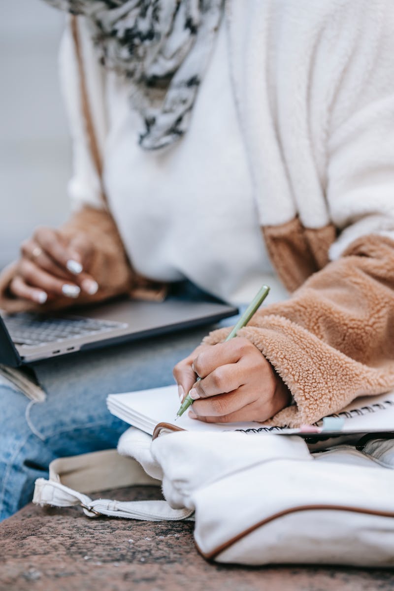 Unrecognizable female student taking notes while preparing for exam using netbook on street