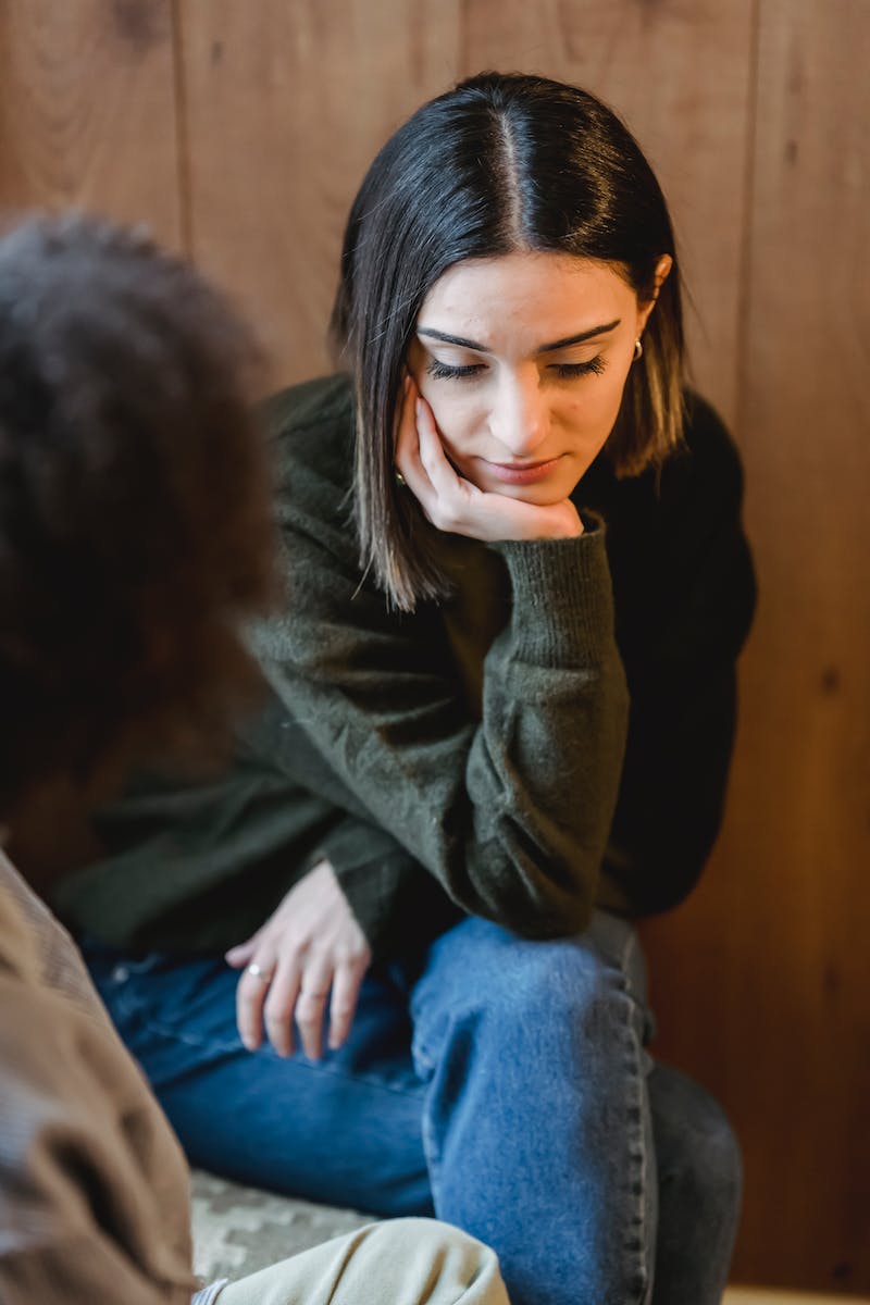 High angle of disappointed young female leaning on hand while sitting with crossed legs near crop girlfriend