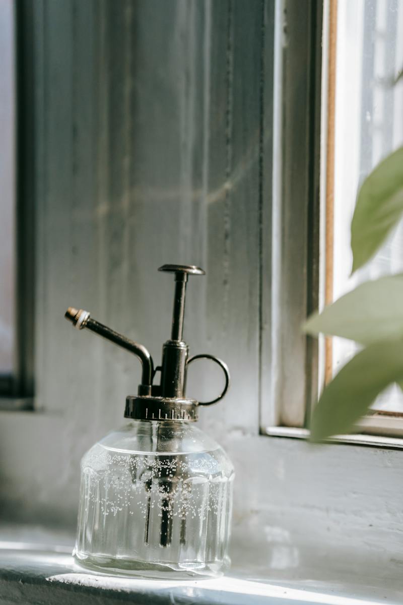 Transparent glass bottle sprayer with fresh water placed on white windowsill in daylight