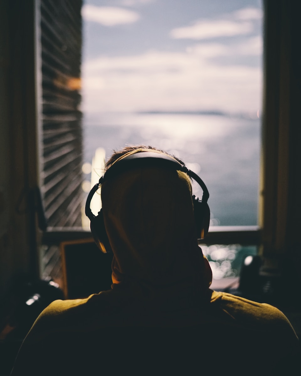 tilt-shift lens photography of person wearing headphones facing body of water through window of a dark room