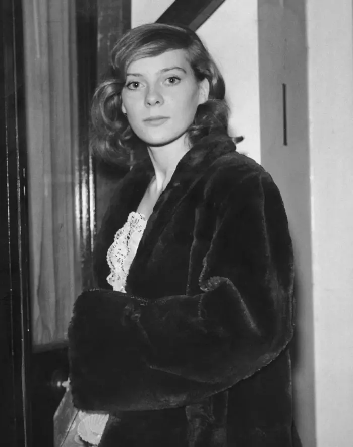 Žena v elegantnom kabáte stojí pri dverách, pohľad smeruje priamo do kamery. Čiernobiele fotografie retro štýlu.