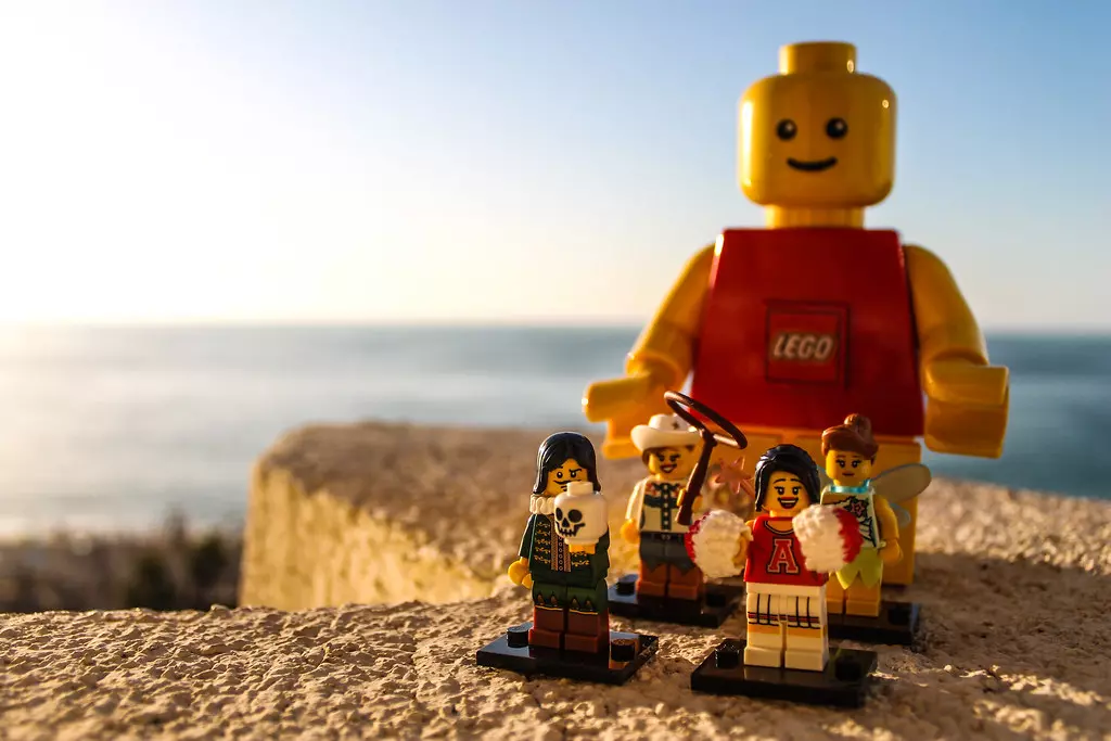 LEGO figúrky a veľká postava na pláži pri zapadajúcom slnku.