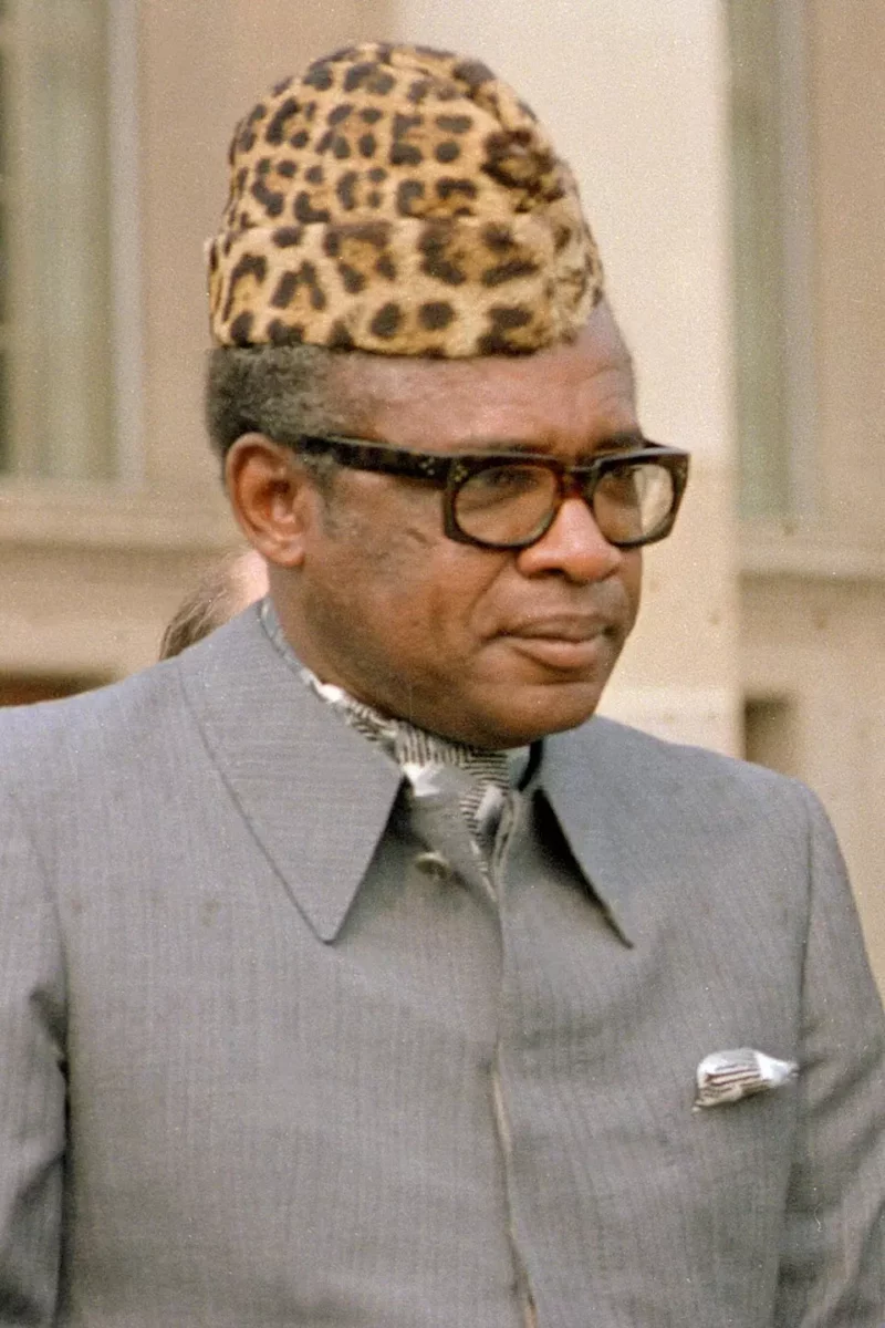 Osoba v obleku a klobúku s leopardím vzorom, pozadie mestská architektúra.