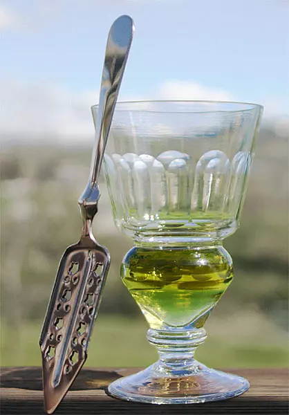 Pohár absintu s lyžičkou na cukor na drevenom stole, rozmazané pozadie vidieka.