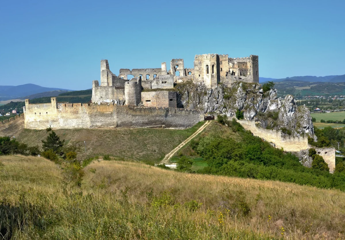 Hrad na skalnatom kopci s výhľadom na zeleň a modrú oblohu. Starobylá architektúra s rozpadnutými múrmi.