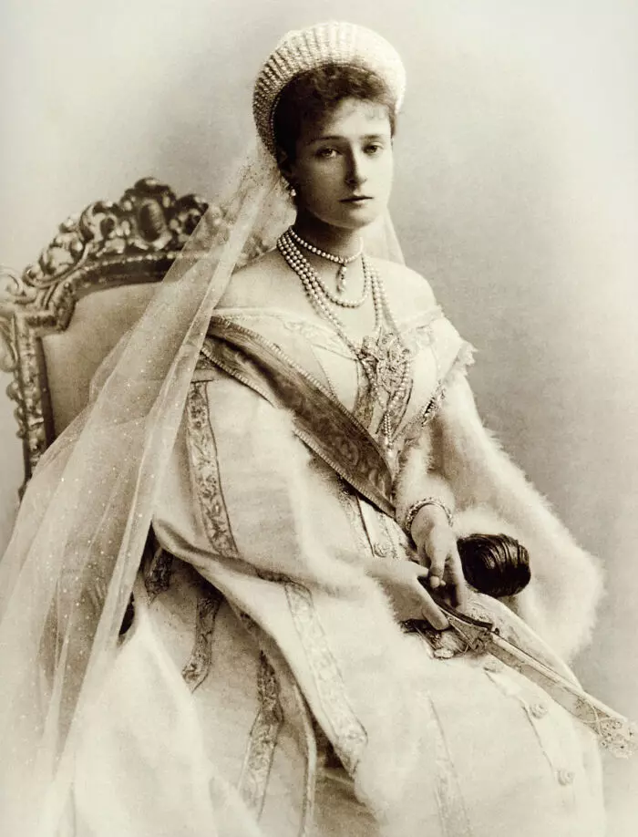 Mladá žena v historickom odeve sedí na zdobenej stoličke, jemný závoj a perly dotvárajú elegantný vzhľad.