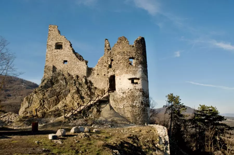 Hradná ruina na kopci za jasného modrého neba, obklopená prírodou a lesmi.