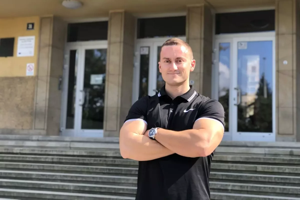 Osoba v čiernom tričku stojí pred budovou so schodmi, ruky má prekrížené, usmieva sa na slnečnom dni.
