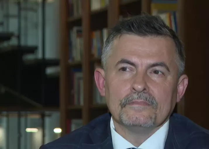 Osoba v obleku pred policou s knihami, pohľad bokom, tlmené svetlo v pozadí.