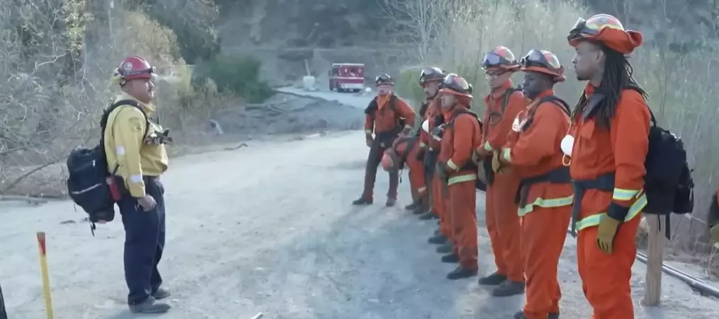 Skupina hasičov v oranžových uniformách pri tréningu v prírode, vedená veliteľom na štrkovitej ceste.
