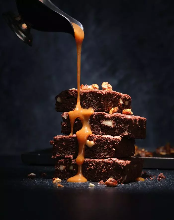 Čokoládové brownies s orechmi a karamelom, pomaly preliate polevou na tmavom pozadí.