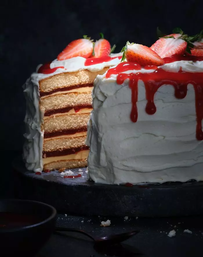 Jahodová torta so šľahačkou a vrstvami biskvitu, ozdobená plátkami jahôd, nakrojená na tmavom pozadí.