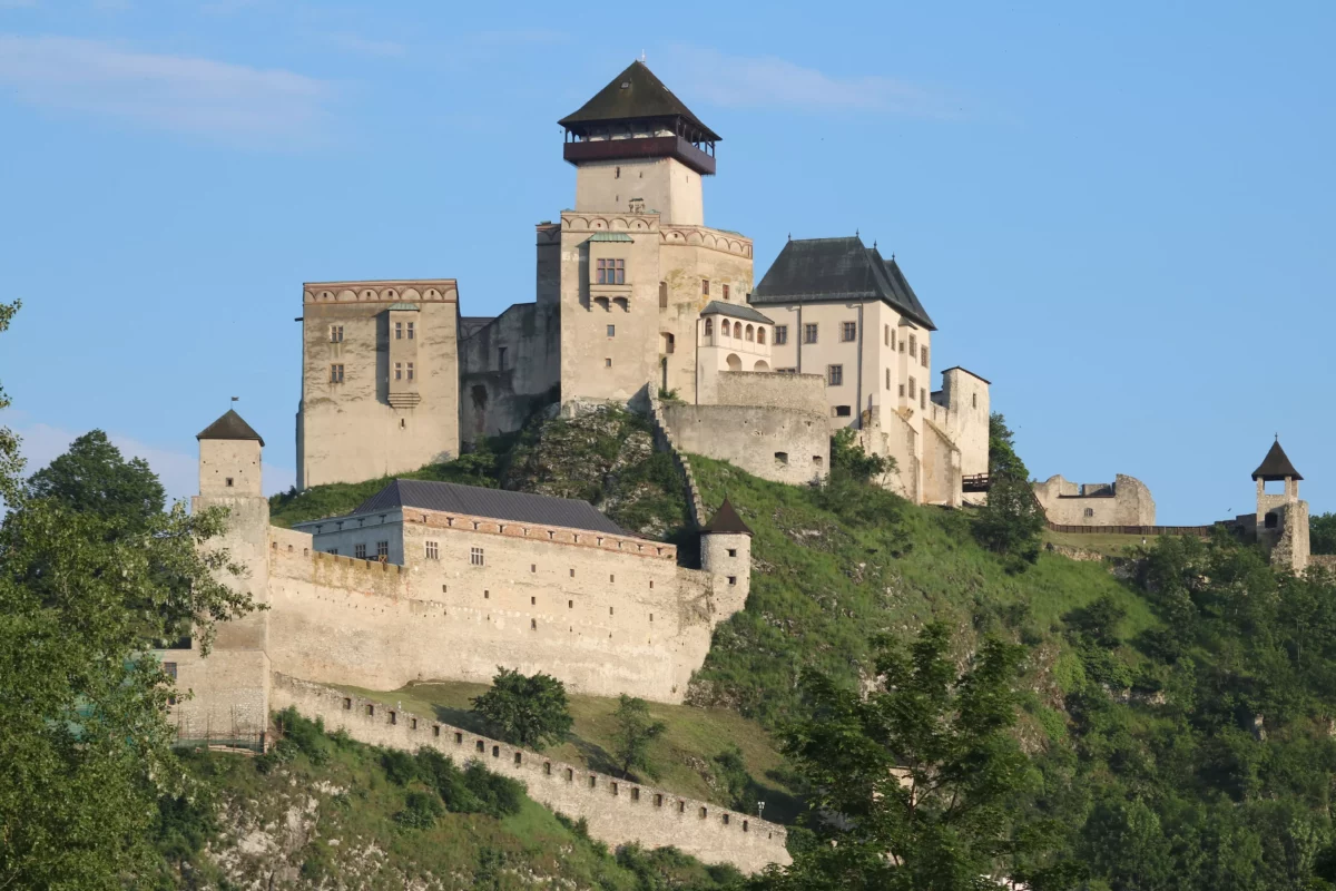 Stredoveký hrad s vežami a hradbami na kopci, obklopený zeleňou a modrou oblohou v pozadí.