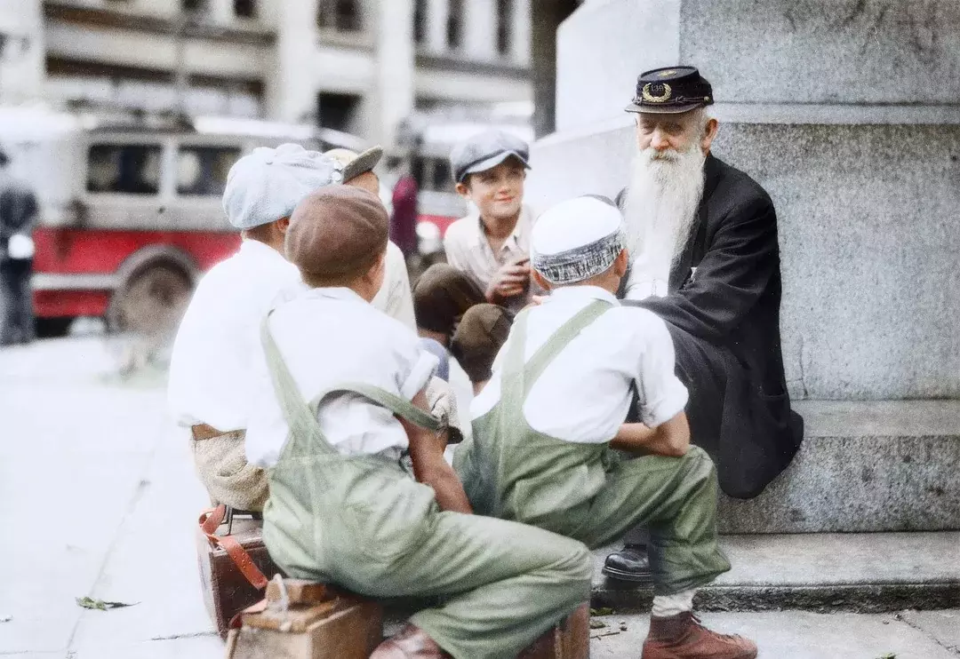 Starší muž rozpráva príbehy skupine detí na ulici; farebná historická fotografia.