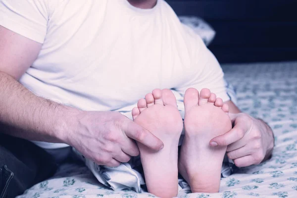 Osoba v bielom tričku sedí na posteli a masíruje si chodidlá, relax a starostlivosť o nohy.