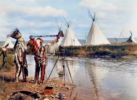 Domorodí Američania pri rieke skúmajú okolie s táborom tipi na pozadí.