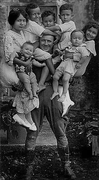 Muž s niekoľkými malými deťmi v náručí pózuje na historickej čiernobielej fotografii.