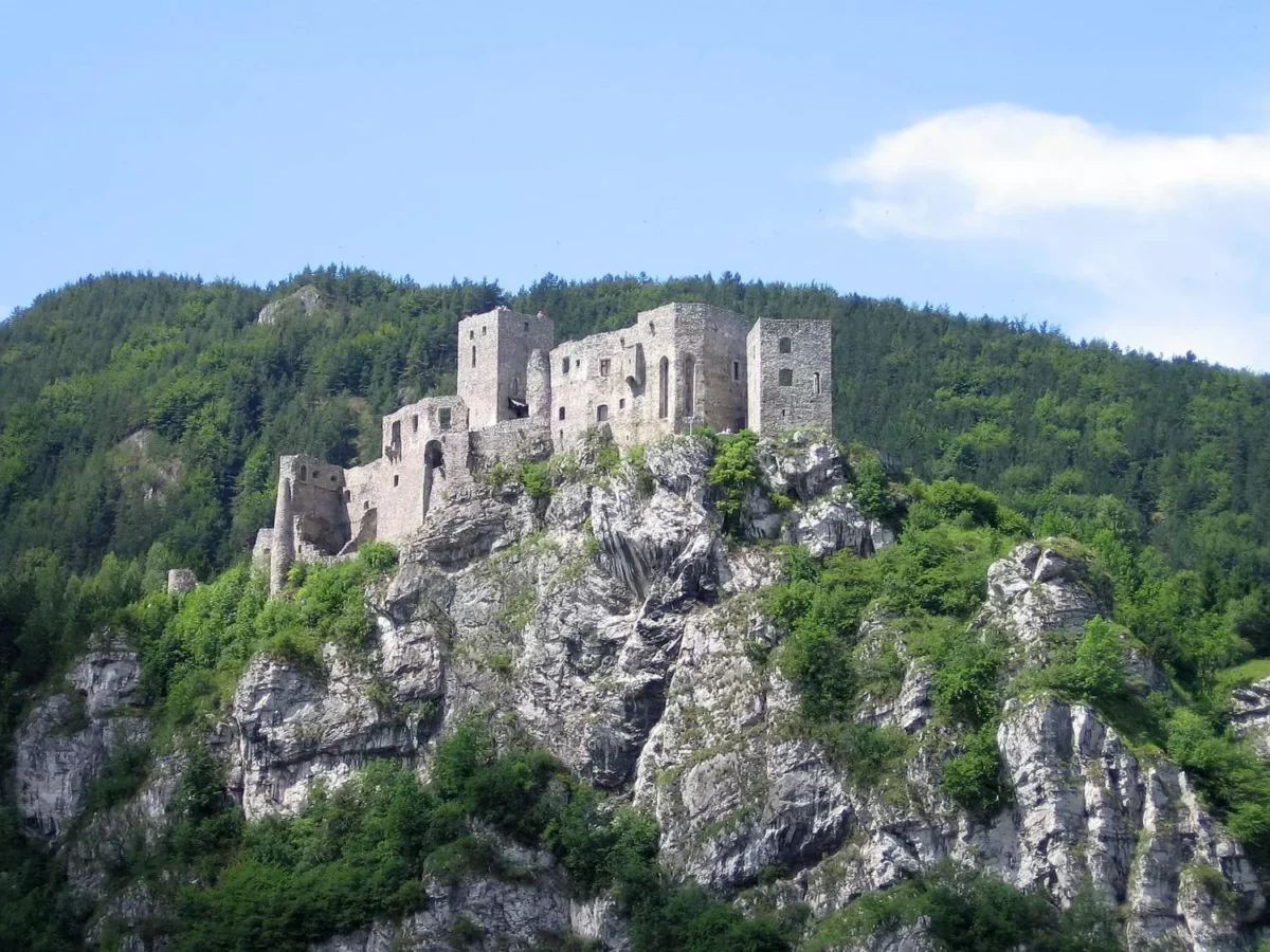 Kamenná zrúcanina hradu na vrchole kopca, obklopená lesmi a modrou oblohou v pozadí.