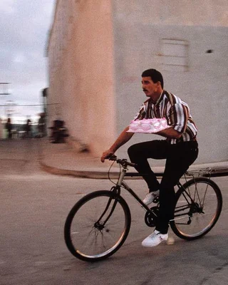 Muž na bicykli s balíčkom v ruke, jazdiaci po mestskom okolí pri súmraku.