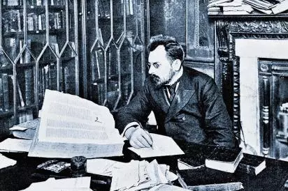 Muž číta veľkú knihu pri stole, obklopený ďalšími knihami a papiermi v knižnici.