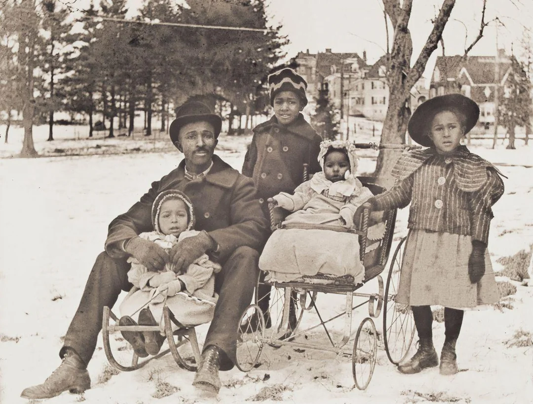 Rodina na zasneženej lúke so sánkami a kočíkom, stará čiernobiela fotografia.