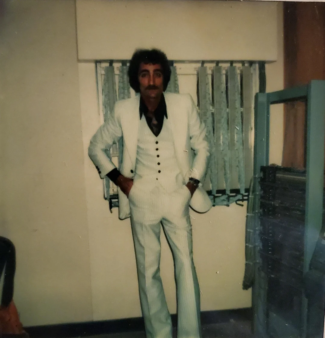 Muž v retro bielom obleku stojí v miestnosti so žalúziami a opiera sa o stenu.