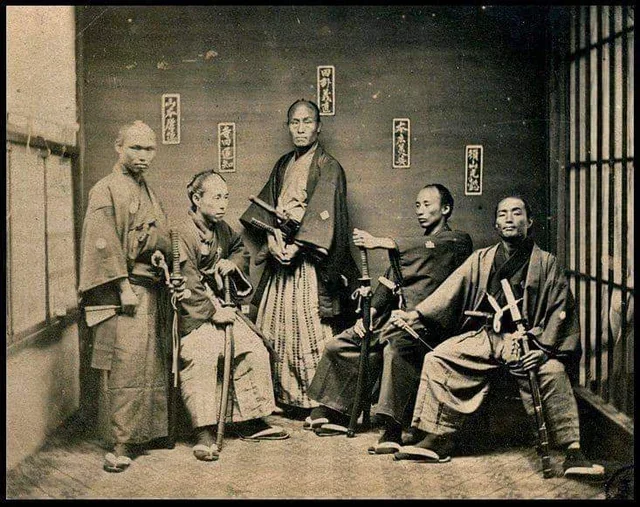 Skupina samurajov v tradičných odevoch, pózuje s mečmi v historickom japonskom interiéri.