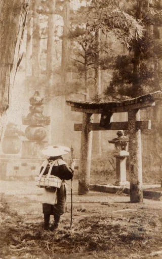 Mních kráča lesom k tradičnej japonskej bráne torii, obklopený starobylými stromami a kamennými sochami.