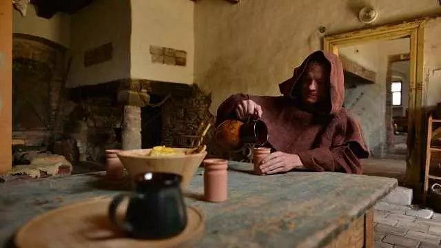 Mních v hnedou kutne nalieva tekutinu do pohára v stredovekej izbe s rustikálnym dreveným stolom.