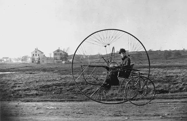 Historická fotografia muža na bicykli s obrovským predným kolesom na vidieckej ceste.