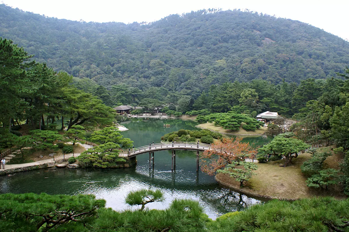 Japonská záhrada s mostom nad rybníkom, obklopená zeleňou a horami, ponúka pokoj a harmonický výhľad.