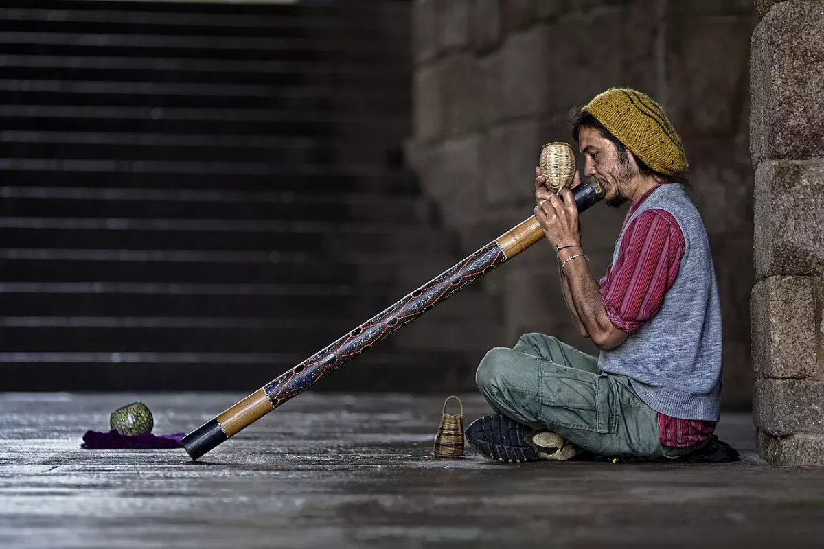 Muž hrá na didgeridoo v tuneli, zatiaľ čo sedí na zemi. Tradičný hudobný nástroj vytvára unikátny zvuk v mestskom prostredí.
