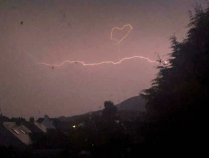 Blesk na nočnej oblohe tvorí tvar srdca nad mestom, v pozadí jemne osvetlené domy.