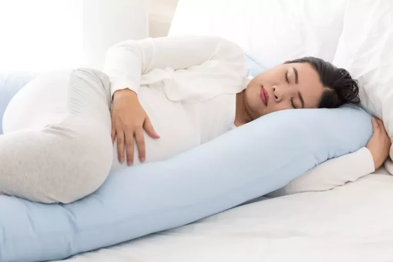 Těhotná žena spí pohodlne na boku s podporným vankúšom v bielej posteli.