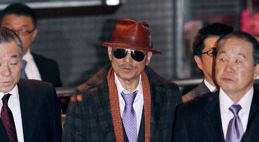 Osoba v klobúku a okuliaroch na verejnom podujatí so skupinou elegantne oblečených ľudí.