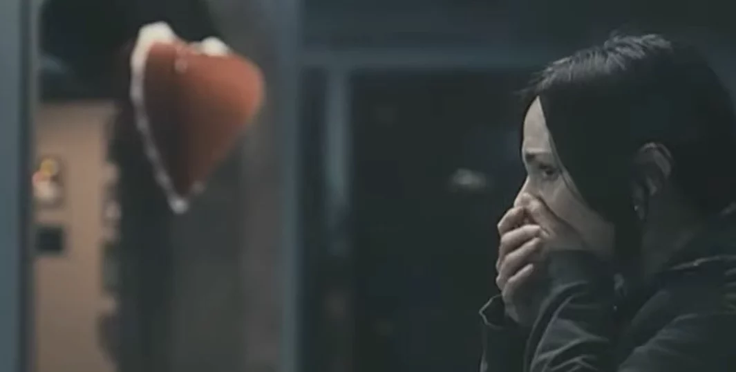 Osoba so zakrytými ústami hľadí na závesné srdce v temnej miestnosti, vyjadruje prekvapenie alebo strach.