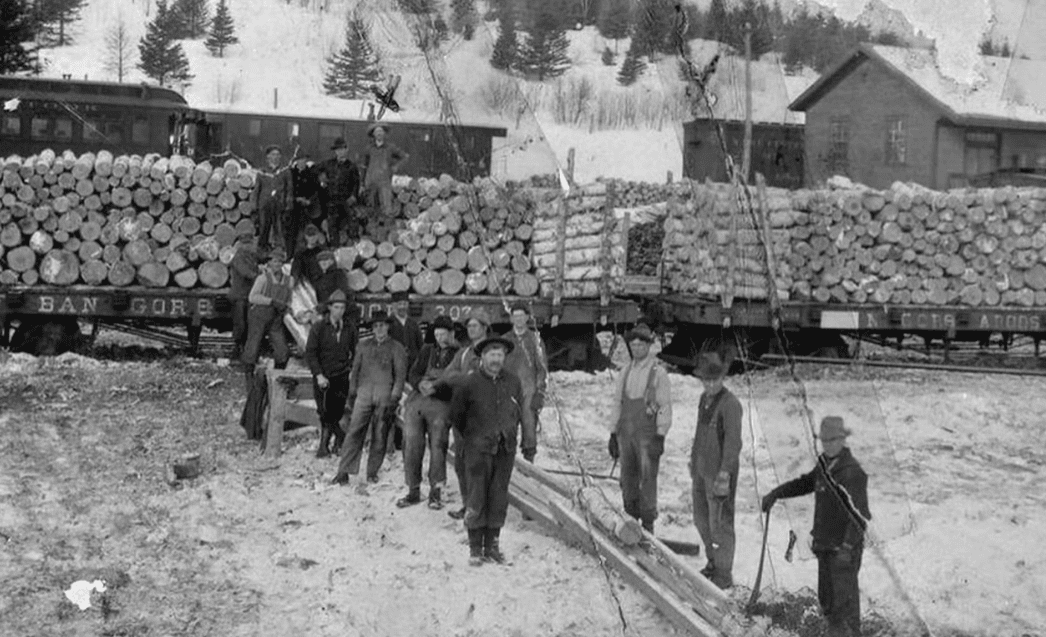 Historická fotografia robotníkov naložených drevom počas zimy, vlak a zasnežená krajina v pozadí.