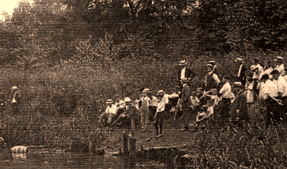 Skupina ľudí na brehu rieky v historických odevoch, rekreácia v prírode, vintage fotografia.