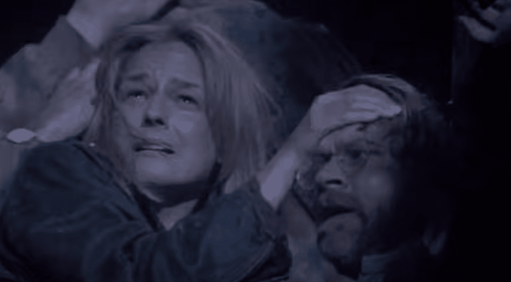 Žena a muž v ustráchanom objatí, zjavne v núdzi, v tmavej scéne so zranením na čele muža.