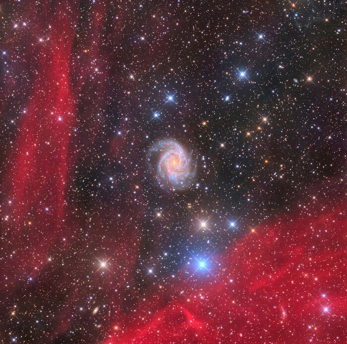 Špirálová galaxia obklopená jasnými hviezdami a červenými prachovými mračnami v hlbokom vesmíre.