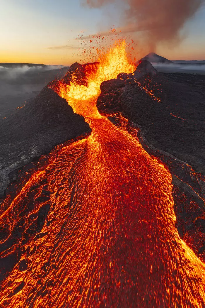 Erupcia sopky s lávovým tokom pri západe slnka, žiariaca oranžovo na tmavom pozadí.