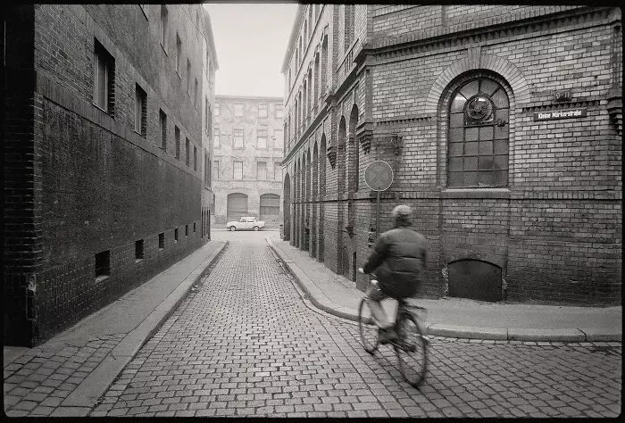 Muž na bicykli prechádza úzkou uličkou so starými tehlovými budovami, v pozadí zaparkované auto.